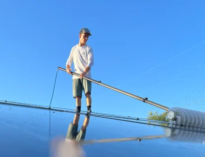 A professional cleaning service worker using a pole to clean a roof under a clear blue sky